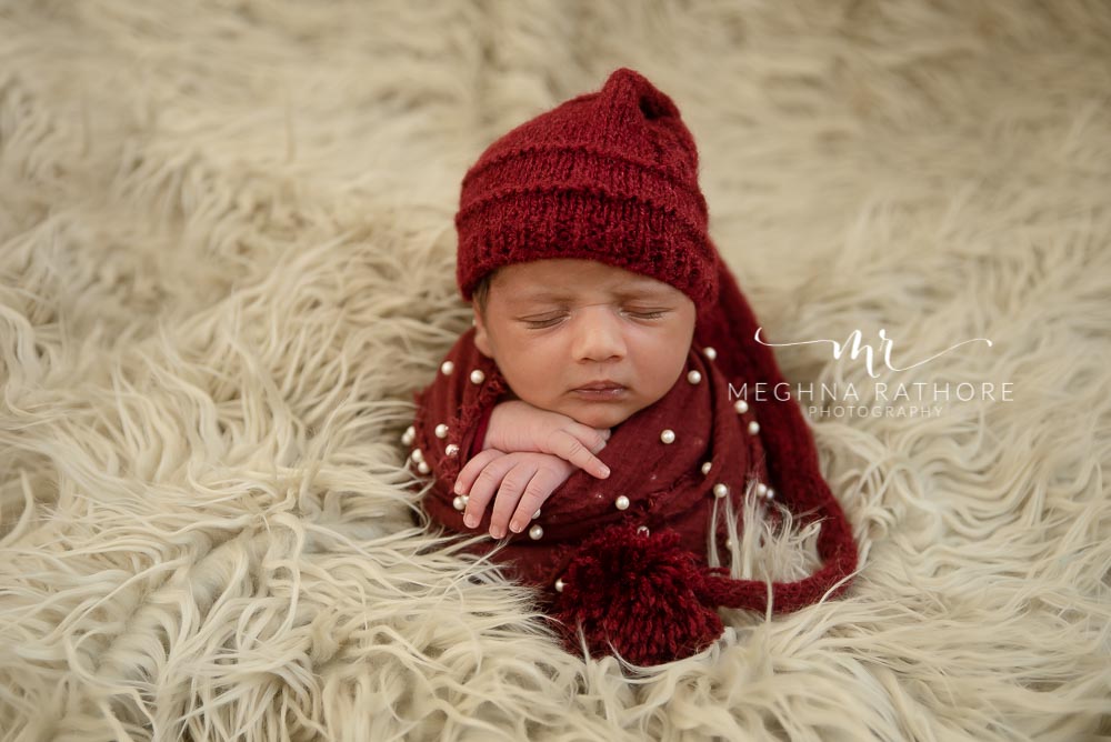 23 days old baby boy wrapped in red cloth and surrounded by white furry blanket at meghna rathore photography in delhi