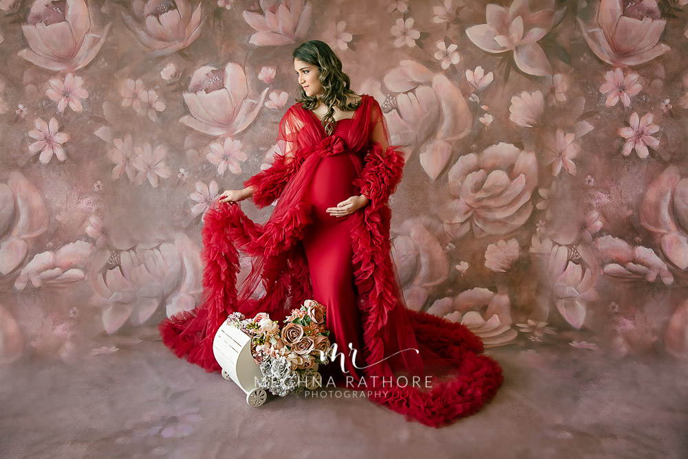 expecting mother wearing red dress small crate with flowers brown backdrop maternity photo shoot meghna rathore photography