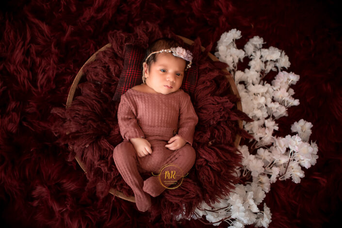 An adorable infant girl in red attire resting on a cozy maroon blanket captured by Delhi NCR's best maternal photographer Meghna Rathore.