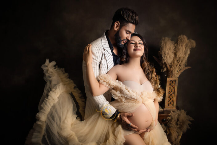 Expectant couple in matching white attire, holding hands and smiling, anticipating the arrival of their baby against a dark background captured by Delhi NCR's best maternal photographer Meghna Rathore.