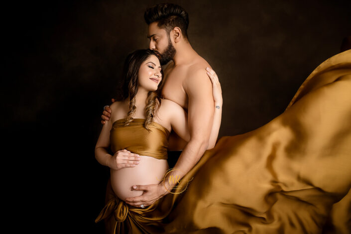 Expectant couple in matching golden attire, holding hands and smiling, anticipating the arrival of their baby captured by Delhi NCR's best maternal photographer Meghna Rathore.