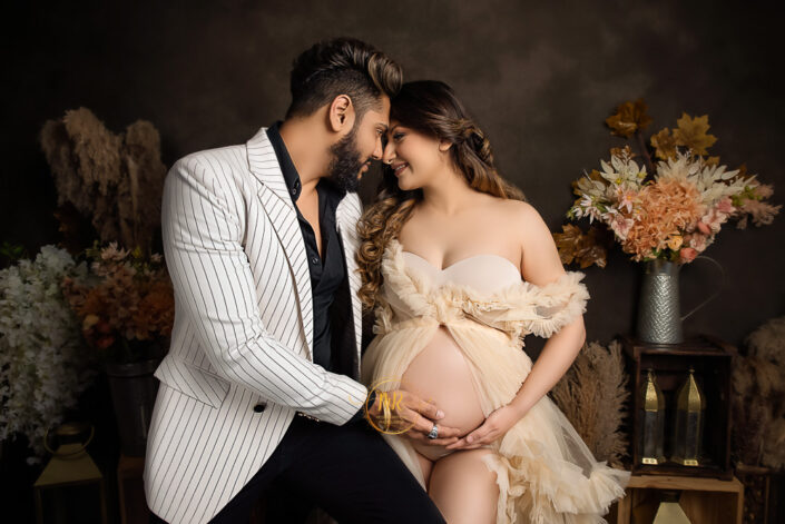 Expectant couple in matching white attire, holding hands and smiling, anticipating the arrival of their baby against a dark background containg flower vase captured by Delhi NCR's best maternal photographer Meghna Rathore.
