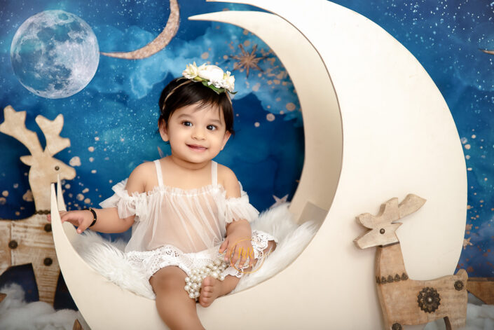 A baby girl in a white dress posing in front of a moon and stars backdrop captured by Delhi NCR's best maternal photographer Meghna Rathore.