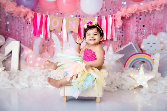 A baby girl in a tutu and rainbow dress happily sits on a white chair captured by Delhi NCR's best maternal photographer Meghna Rathore.