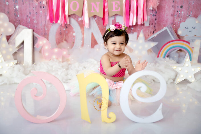 A baby girl in a pink outfit sits in front of a rainbow and a word "one" captured by Delhi NCR's best maternal photographer Meghna Rathore.