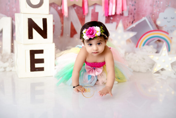 A baby girl in a pink tutu and rainbow skirt sitting on a pink background captured by Delhi NCR's best maternal photographer Meghna Rathore.