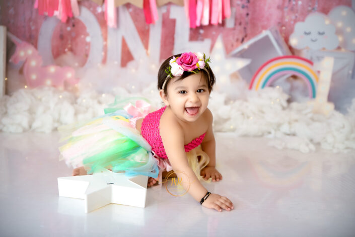 A baby girl in a pink tutu and rainbow skirt sitting on a pink background captured by Delhi NCR's best maternal photographer Meghna Rathore.