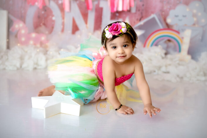 A baby girl in a pink tutu and rainbow skirt sitting on a pink background captured by Delhi NCR's best maternal photographer Meghna Rathore.