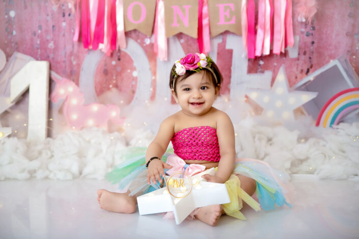 A baby girl in a pink tutu and rainbow skirt sitting on a pink background captured by Delhi NCR's best maternal photographer Meghna Rathore.