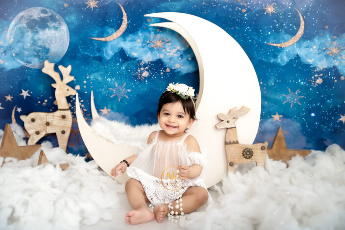 A baby girl in a white dress posing in front of a moon and stars backdrop captured by Delhi NCR's best maternal photographer Meghna Rathore.