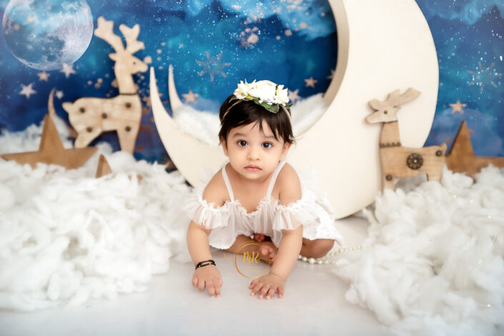 A baby girl in a white dress posing in front of a moon and stars backdrop captured by Delhi NCR's best maternal photographer Meghna Rathore.