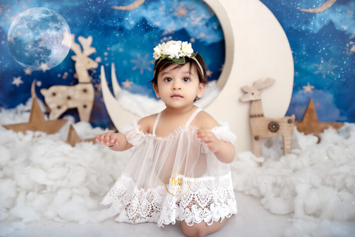 A baby girl in a white dress posing in front of a moon and stars backdrop captured by Delhi NCR's best maternal photographer Meghna Rathore.