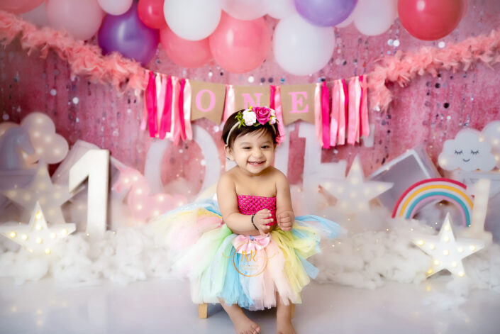 A baby girl in a tutu and rainbow dress happily sits on a white chair captured by Delhi NCR's best maternal photographer Meghna Rathore.