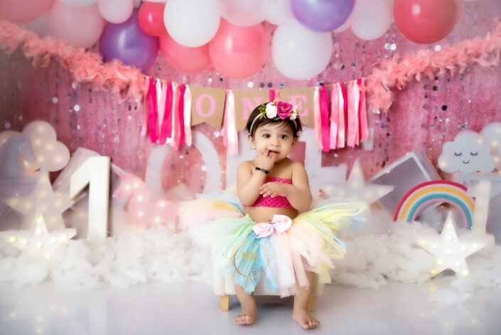 A baby girl in a tutu and rainbow dress happily sits on a white chair captured by Delhi NCR's best maternal photographer Meghna Rathore.