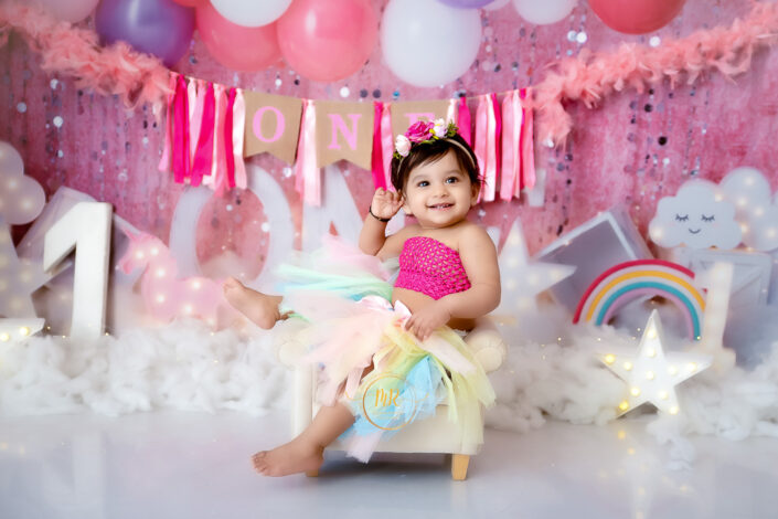 A baby girl in a tutu and rainbow dress happily sits on a white chair captured by Delhi NCR's best maternal photographer Meghna Rathore.