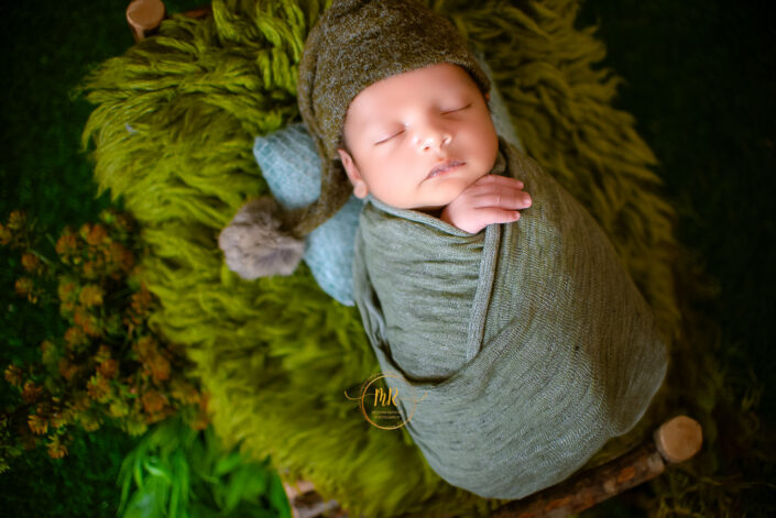 Newborn baby boy wrapped in green blanket lying on grassy field captured by Delhi NCR's best maternal photographer Meghna Rathore.