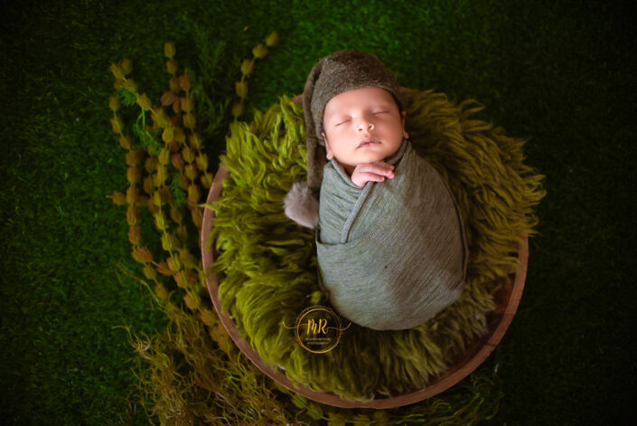 Newborn baby boy wrapped in green blanket lying on grassy field captured by Delhi NCR's best maternal photographer Meghna Rathore.