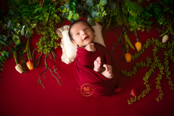 A baby in a red wrap laying on a red background captured by Delhi NCR's best maternal photographer Meghna Rathore.