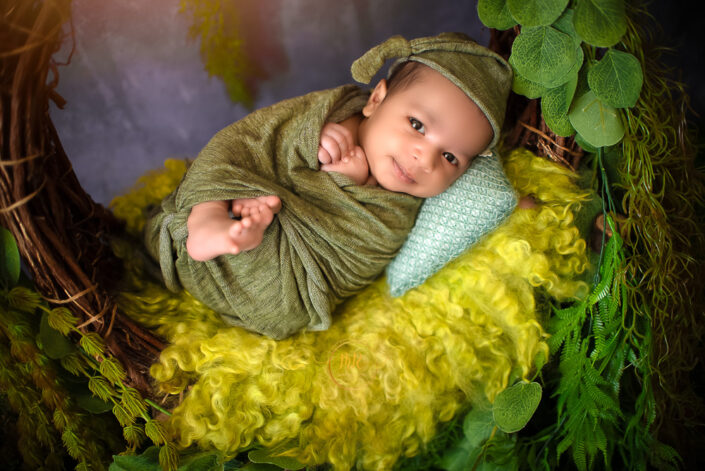 A newborn baby boy wrapped in a green blanket sitting in a wooden basket captured by Delhi NCR's best maternal photographer Meghna Rathore.