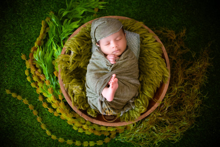 A newborn baby boy wrapped in a green blanket sitting in a wooden bowl captured by Delhi NCR's best maternal photographer Meghna Rathore.