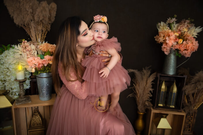A woman in a pink dress cradling a baby in her arms, radiating love and tenderness with flower vases around captured by Delhi NCR's best maternal photographer Meghna Rathore.