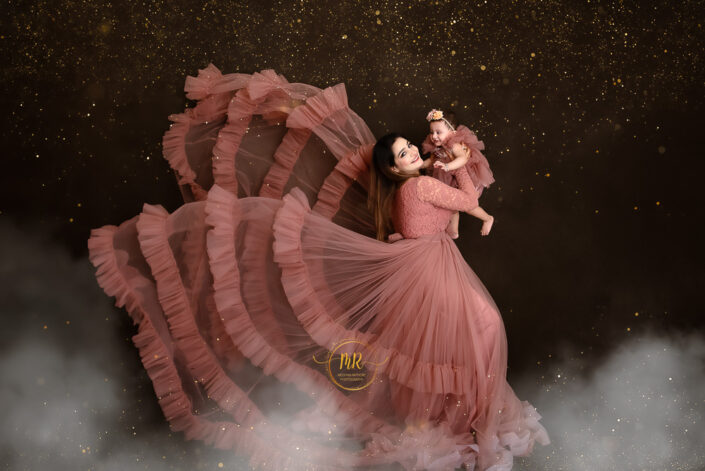 Woman in pink dress holding her baby, smiling warmly against a gliterry background captured by Delhi NCR's best maternal photographer Meghna Rathore.