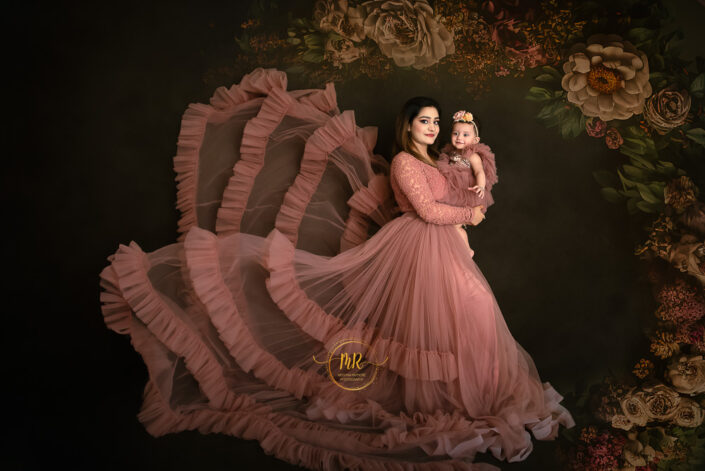 Woman in pink dress holding her baby, smiling warmly against a floral backdrop captured by Delhi NCR's best maternal photographer Meghna Rathore.