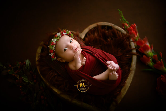 A baby girl in a red dress smiling indide a heart shaped basket captured by Delhi NCR's best maternal photographer Meghna Rathore.