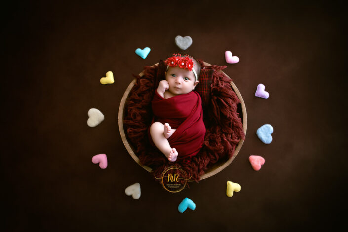 A baby girl in a red dress smiling, surrounded by hearts captured by Delhi NCR's best maternal photographer Meghna Rathore.