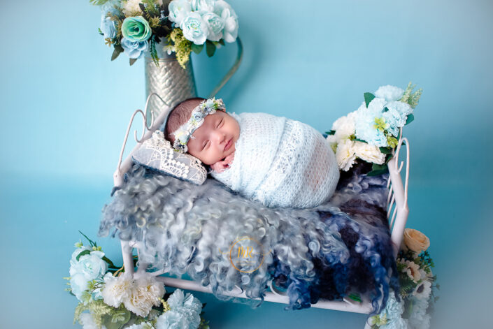 A newborn girl wrapped in a blue blanket, surrounded by flowers captured by Delhi NCR's best maternal photographer Meghna Rathore.
