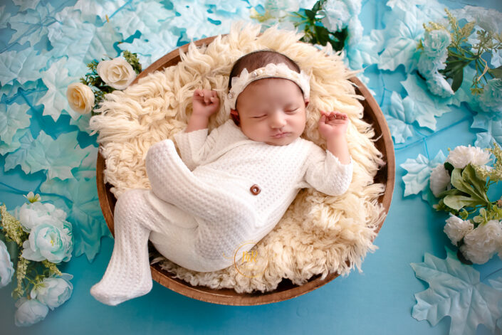 A newborn baby girl peacefully sleeping in a basket on a blue background captured by Delhi NCR's best maternal photographer Meghna Rathore.