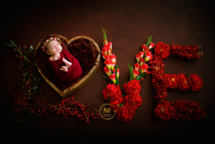 A baby girl in a red dress felling sleeping indide a heart shaped basket ,forming the word "Love" captured by Delhi NCR's best maternal photographer Meghna Rathore.