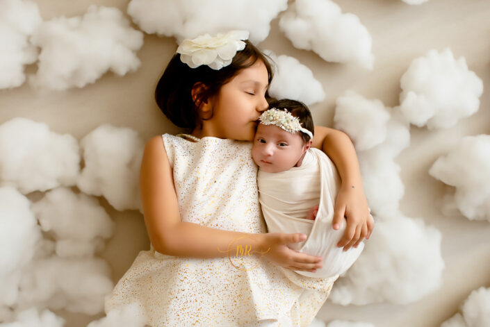 A little girl with her newborn sibling relaxing on a soft white fur rug, enjoying a playful moment together moment together captured by Delhi NCR's best maternal photographer Meghna Rathore.