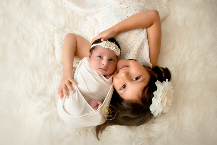 A little girl with her newborn sibling relaxing on a soft white fur rug, enjoying a playful moment together moment together captured by Delhi NCR's best maternal photographer Meghna Rathore.