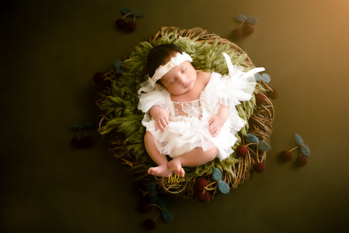 A newborn girl in a white dress sitting among cherries captured by Delhi NCR's best maternal photographer Meghna Rathore.
