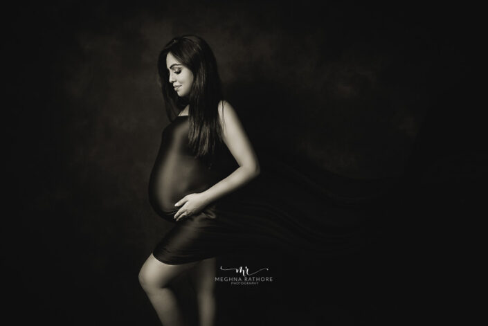 A pregnant woman in a stylish black dress posing for a black and white photo captured by Delhi NCR's best maternal photographer Meghna Rathore.