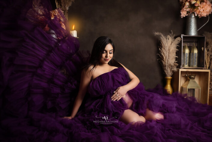 A pregnant woman in a purple dress sitting on a bed captured by Delhi NCR's best maternal photographer Meghna Rathore.