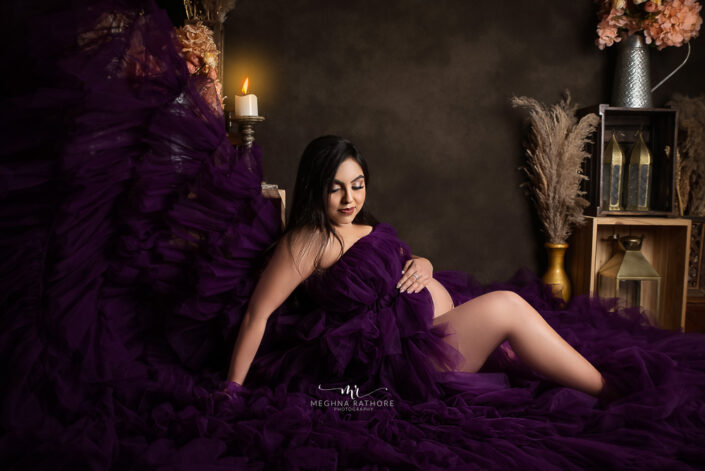 A pregnant woman in a purple dress sitting on a bed captured by Delhi NCR's best maternal photographer Meghna Rathore.