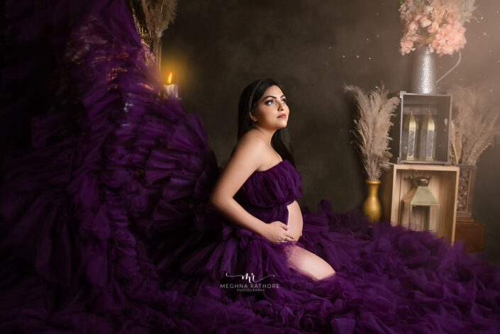 A pregnant woman in a purple dress sitting on a bed captured by Delhi NCR's best maternal photographer Meghna Rathore.