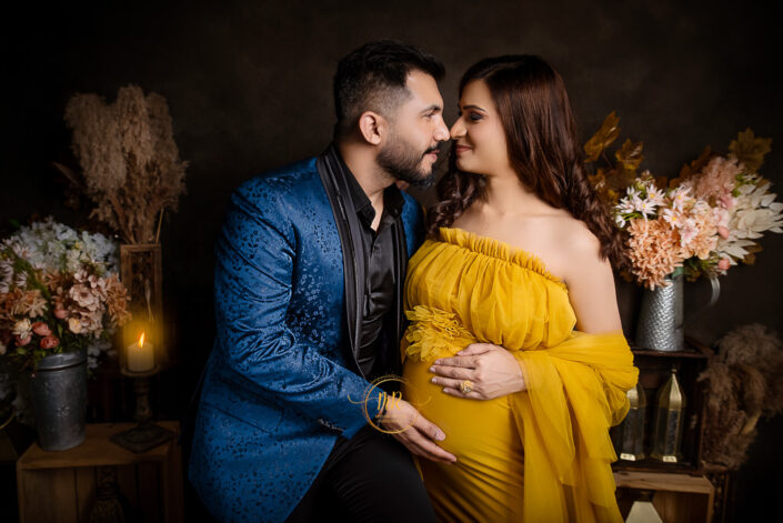 An expectant couple in elegant yellow and blue outfits posing with flowers and a candle in the background captured by Delhi NCR's best maternal photographer Meghna Rathore.