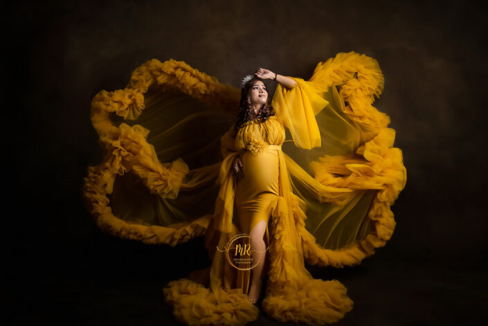 A pregnant woman in yellow dress smiling for a photo against a brown backdrop captured by Delhi NCR's best maternal photographer Meghna Rathore.