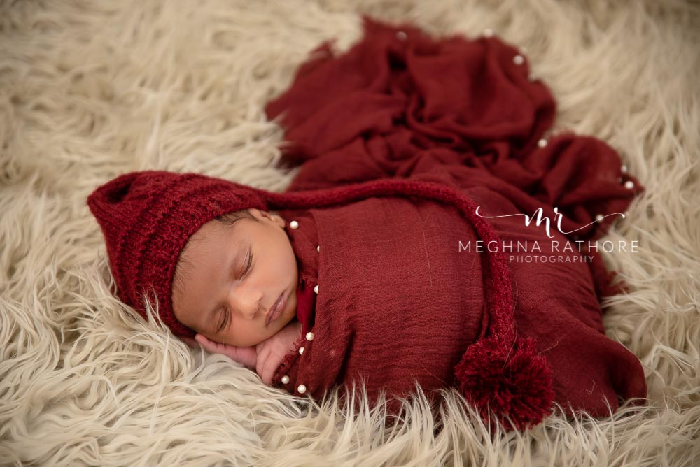 23 days old baby boy tucked in red colored cloth and woolen cap in a furry white blanket and posing at meghna rathore photography in delhi