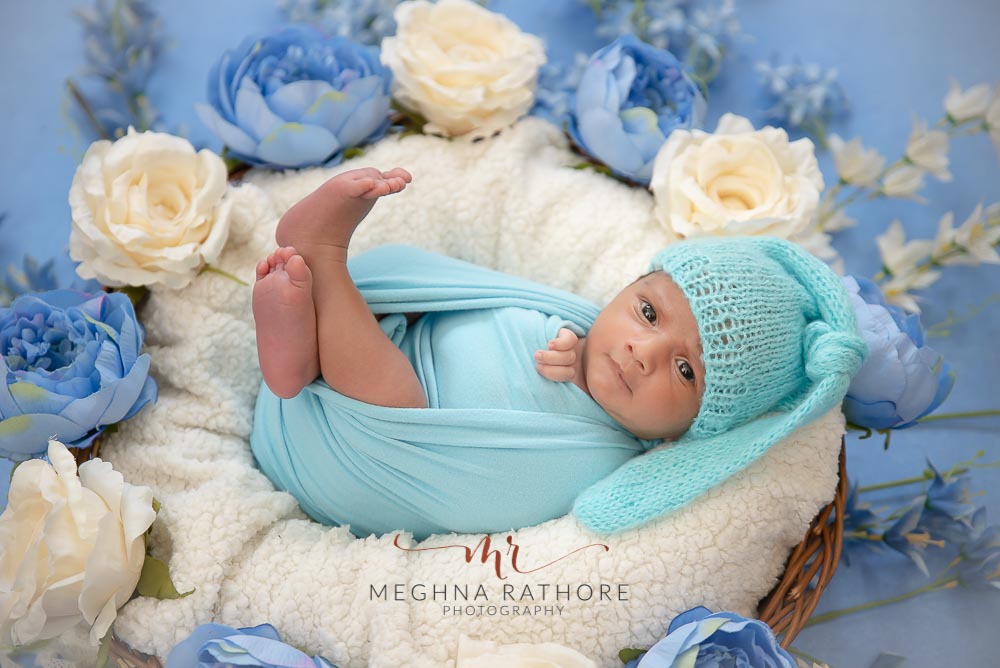 23 days old newborn baby boy tucked in a white blanket covered basket with white and blue flowers around him and posing at meghna rathore photography in delhi
