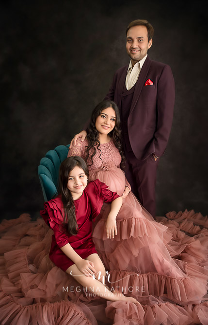 Professional shoot of Mother-to-be with partner and daughter in peach gown, red dress and purple suit during maternity shoot with black background in Delhi