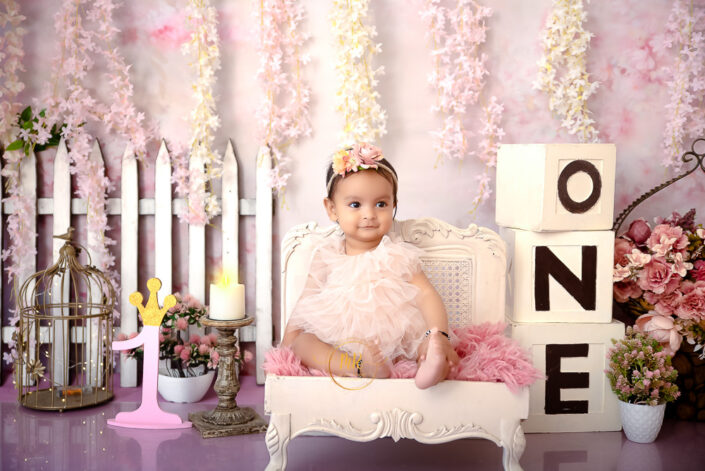 An adorable infant girl sitting on a white couch against pink backdrop with the number "one" displayed prominently captured by Delhi NCR's best maternal photographer Meghna Rathore.
