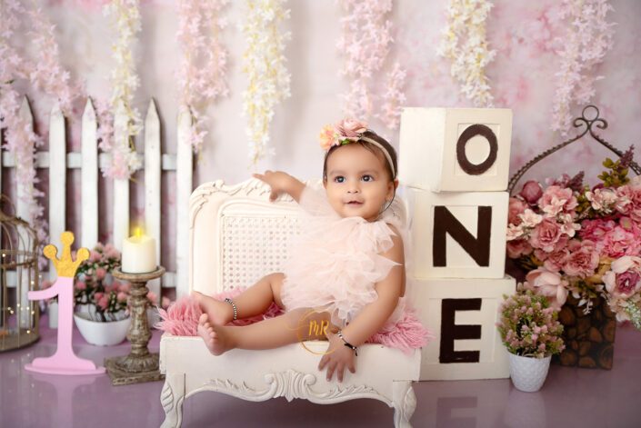 An adorable infant girl sitting on a white couch against pink backdrop with the number "one" displayed prominently captured by Delhi NCR's best maternal photographer Meghna Rathore.