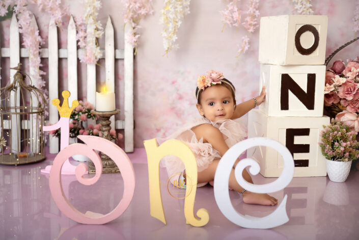 An adorable infant girl on a pink backdrop with the number "one" displayed prominently captured by Delhi NCR's best maternal photographer Meghna Rathore.