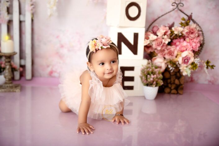 A baby girl in a pink dress playing with three blocks on the floor saying "one" captured by Delhi NCR's best maternal photographer Meghna Rathore.