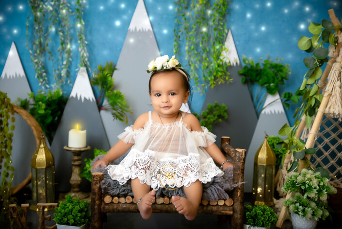 A baby girl in a white dress sitting on a wooden bed with plants and a mountain backdrop captured by Delhi NCR's best maternal photographer Meghna Rathore.