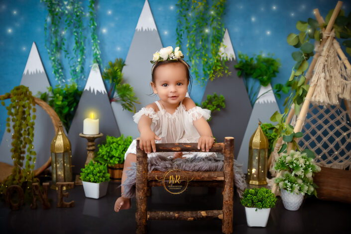 A baby girl in a white dress sitting on a wooden bed with plants and a mountain backdrop captured by Delhi NCR's best maternal photographer Meghna Rathore.